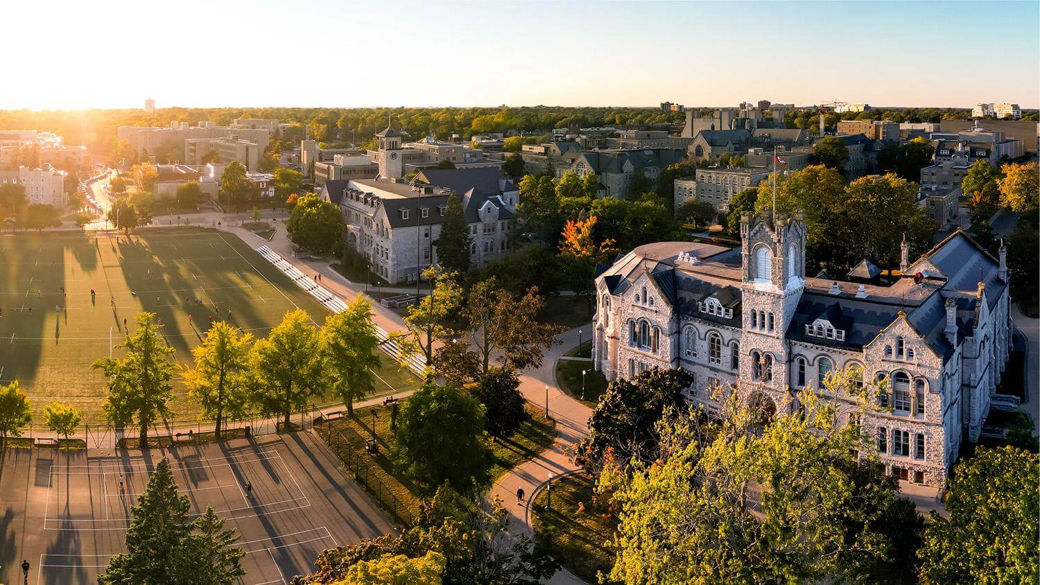 Aerial view of a stone Queen's University building near a green sports field, surrounded by trees and other campus buildings at sunset.