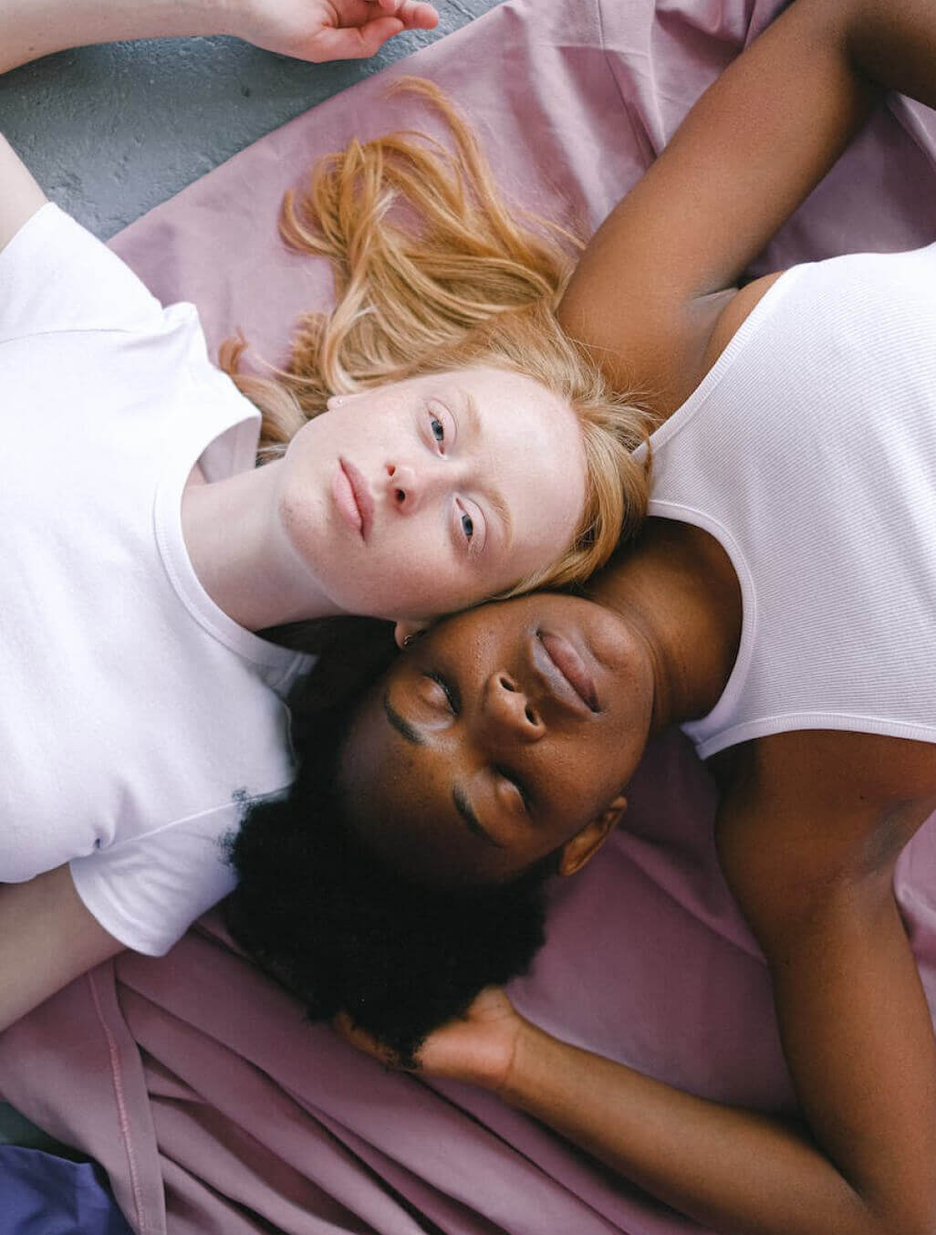 Two people lie on a pink sheet, head to head. One person with light skin and red hair looks up, wearing a white top. The other person with dark skin and black hair rests their head on the others arm, eyes closed, also wearing a white top.