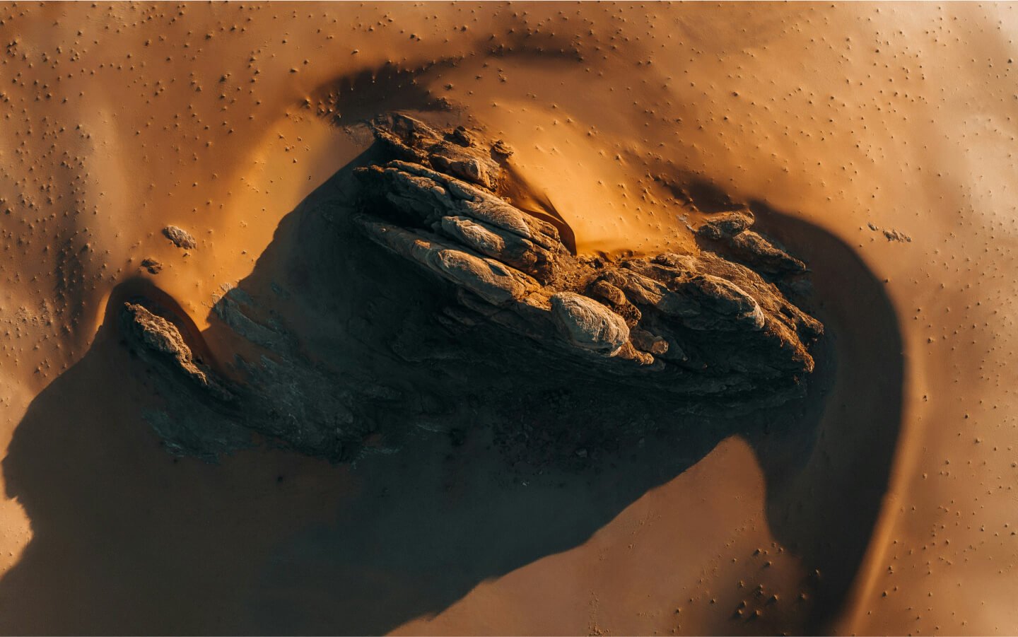 Aerial view of a large rock formation casts a long shadow on smooth, sandy terrain. The earthy tones and shadows create a dramatic contrast, showcasing nature’s impeccable marketing as rugged textures of the rock stand out against the sleek sand.