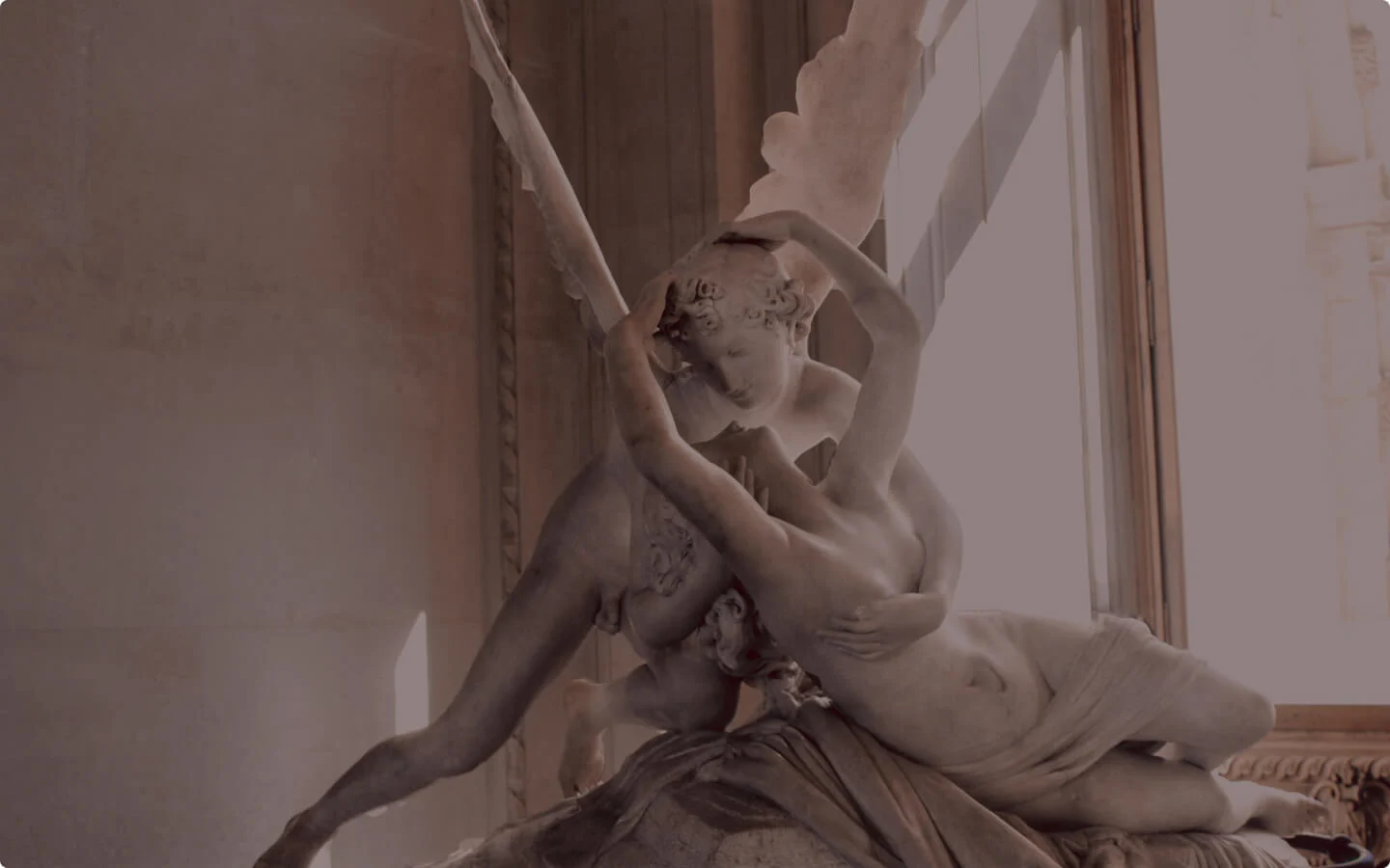In a softly lit museum space near NYU, a marble sculpture depicts two figures in a tender embrace. One, with wings, leans over the other, who reclines elegantly against the backdrop of large windows.