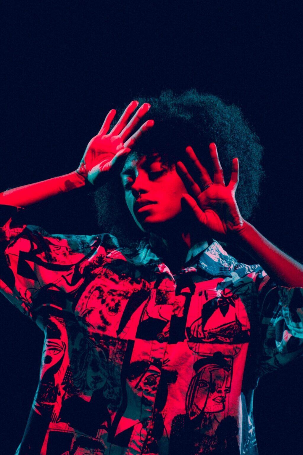 A person with curly hair poses dramatically, their hands near their face, illuminated by red and blue lighting. They wear a patterned shirt featuring abstract designs, evoking an entertainment vibe. The dark background accentuates the vibrant colors and theatrical flair.