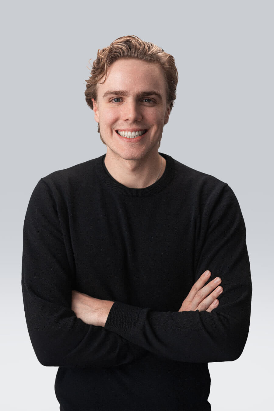 A person with light hair smiles while standing with arms crossed, wearing a black sweater. The background is a gradient from gray to white.