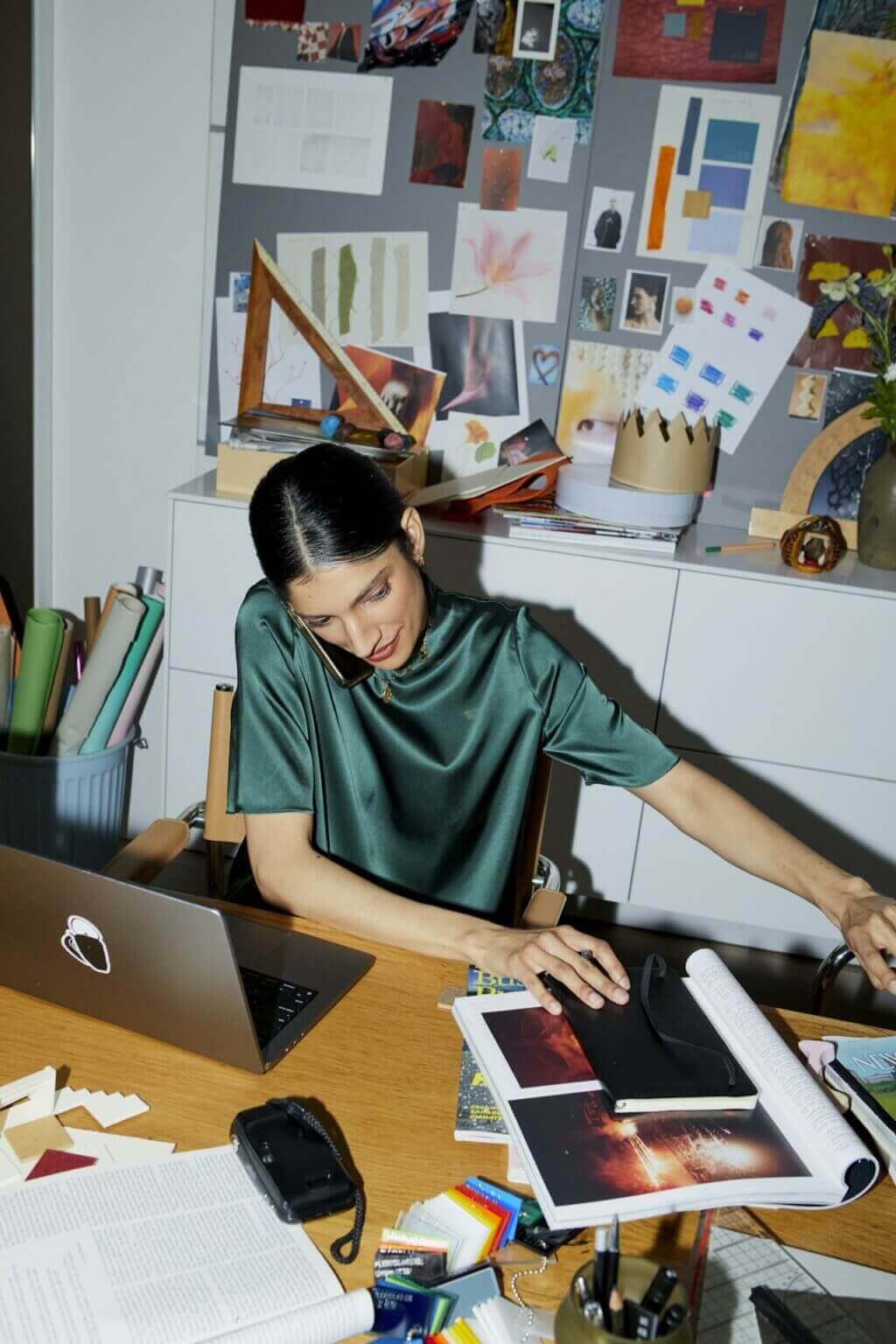 A woman in a satin top sits at a cluttered desk, reaching for an item while brainstorming her next SaaS project. The desk is scattered with a laptop, open magazines, colored pencils, and a camera. Behind her, a board brims with photos and artwork, creating an inspiring artistic workspace.