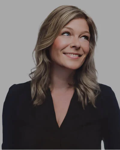 Lindsay Smith, with shoulder-length wavy blonde hair, smiles while looking upwards. Clad in a black top against a plain gray background, their cheerful demeanor radiates warmth and positivity.