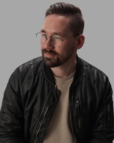 Man with short hair and a beard, wearing round glasses, a black leather jacket, and a beige ribbed shirt, poses against a gray background.