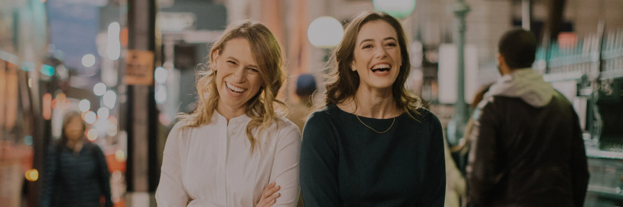 Two women are smiling and laughing while standing on a city street at night. They are casually dressed, and the blurred background shows urban elements like streetlights and pedestrians.