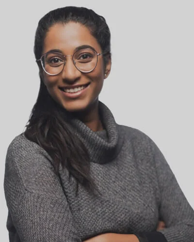 A person with long dark hair and glasses, smiling, wearing a gray turtleneck sweater, stands against a plain light background with arms crossed.