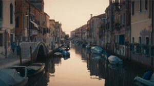 A tranquil canal scene at sunset, bordered by historic buildings, unfolds with the takt of gentle waves. Several small boats float on the calm water, while a stone bridge spans the canal in the distance under a softly lit sky.