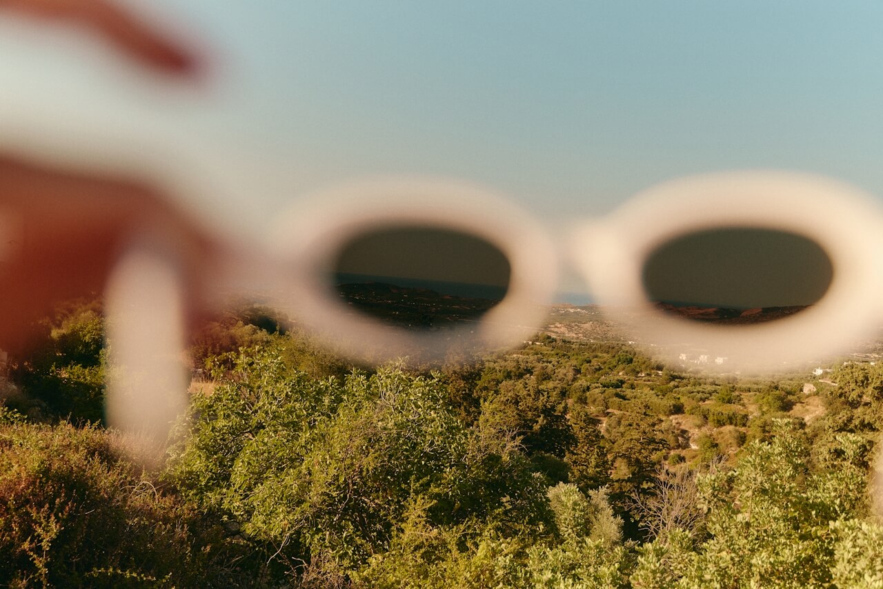 Close-up of a hand holding white sunglasses, focusing through the lenses on a landscape with lush green trees and hills under a clear blue sky, creating a contrast between the blurred and sharp areas.