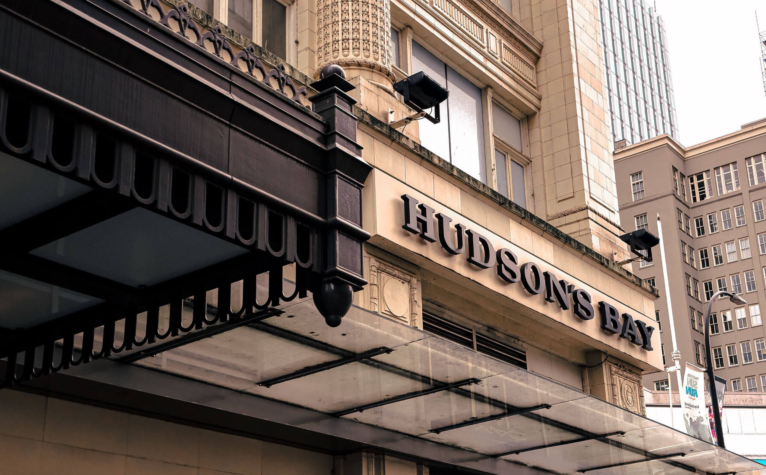 The image shows the entrance to a Hudsons Bay department store. The building features classic architectural details with large windows. The store name is prominently displayed on the facade above a glass awning.