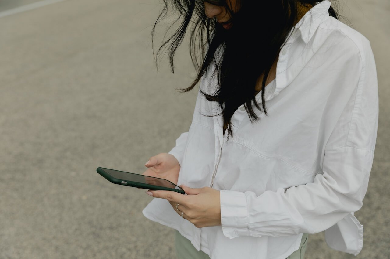 A person with long dark hair wearing a white shirt stands outdoors on a paved surface, looking down and using a smartphone.