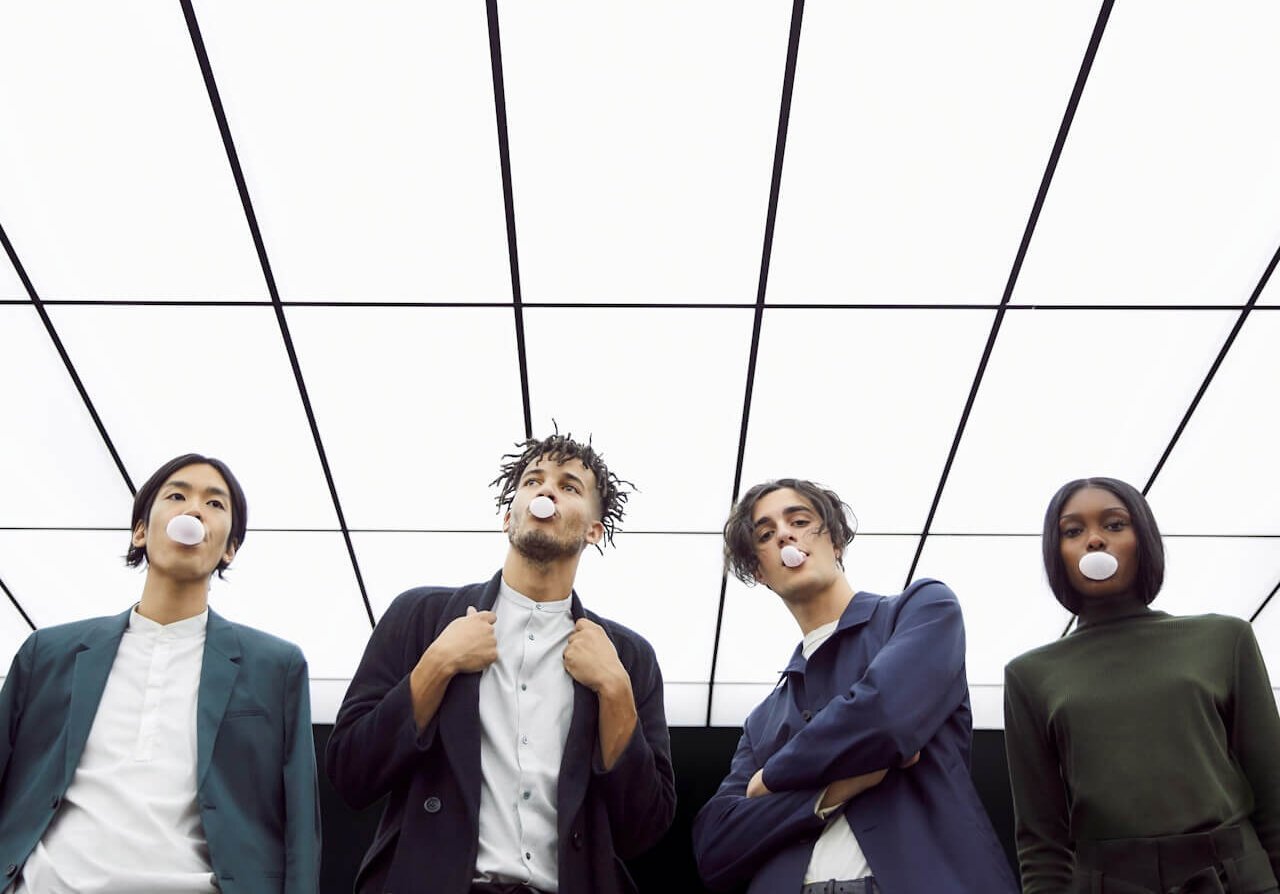Four people are standing under a checkered ceiling, blowing bubbles with gum. They are dressed in stylish, modern clothing, giving off a cool and confident vibe. The perspective is from below, emphasizing the ceiling pattern.