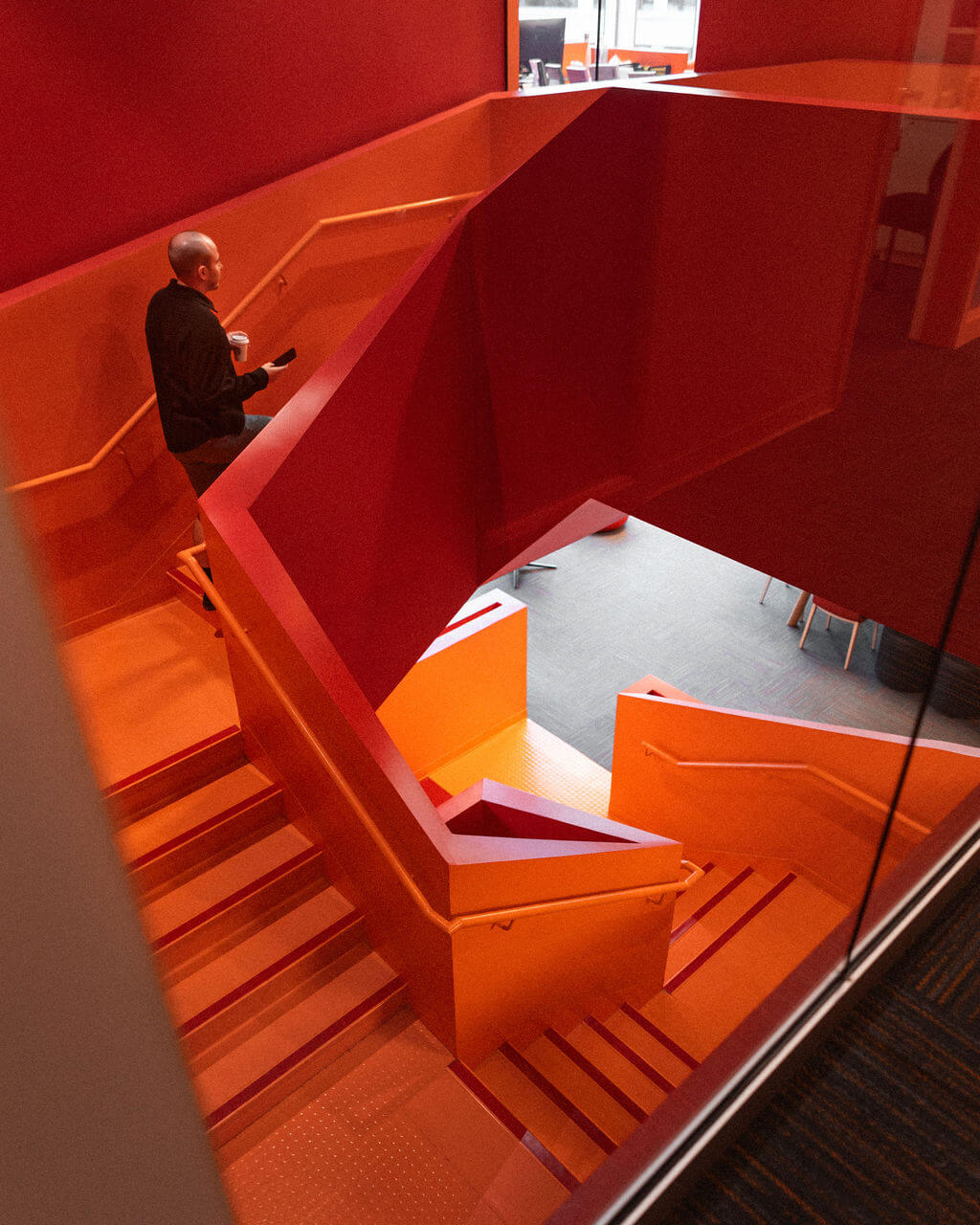 A person ascends bright orange and red modern stairs inside an NYU building, the vivid colors creating bold geometric patterns. The individual is holding something, and the well-lit area features some chairs visible below.