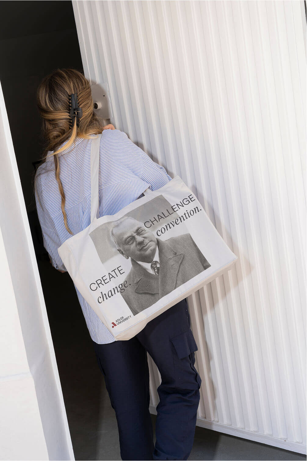 A woman with long hair in a clip carries a tote bag featuring a black-and-white portrait of a man and the text CREATE change. CHALLENGE convention. as she walks through a white NYU doorway.