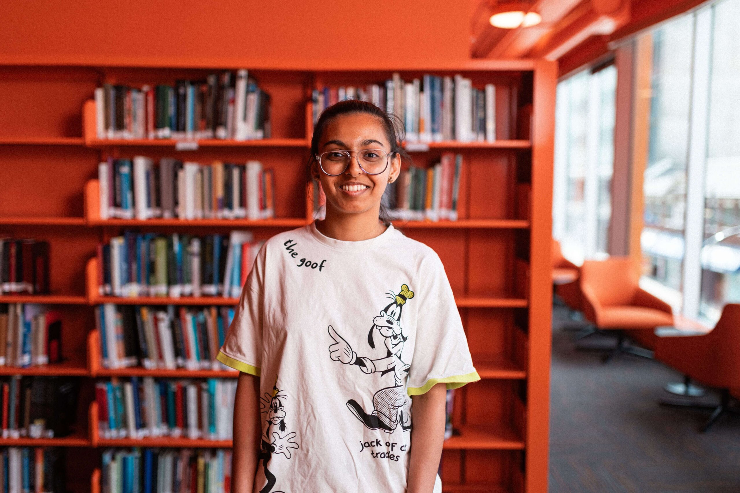 A young woman with glasses and a cartoon t-shirt stands smiling in front of orange bookshelves filled with books in an NYU library with large windows.