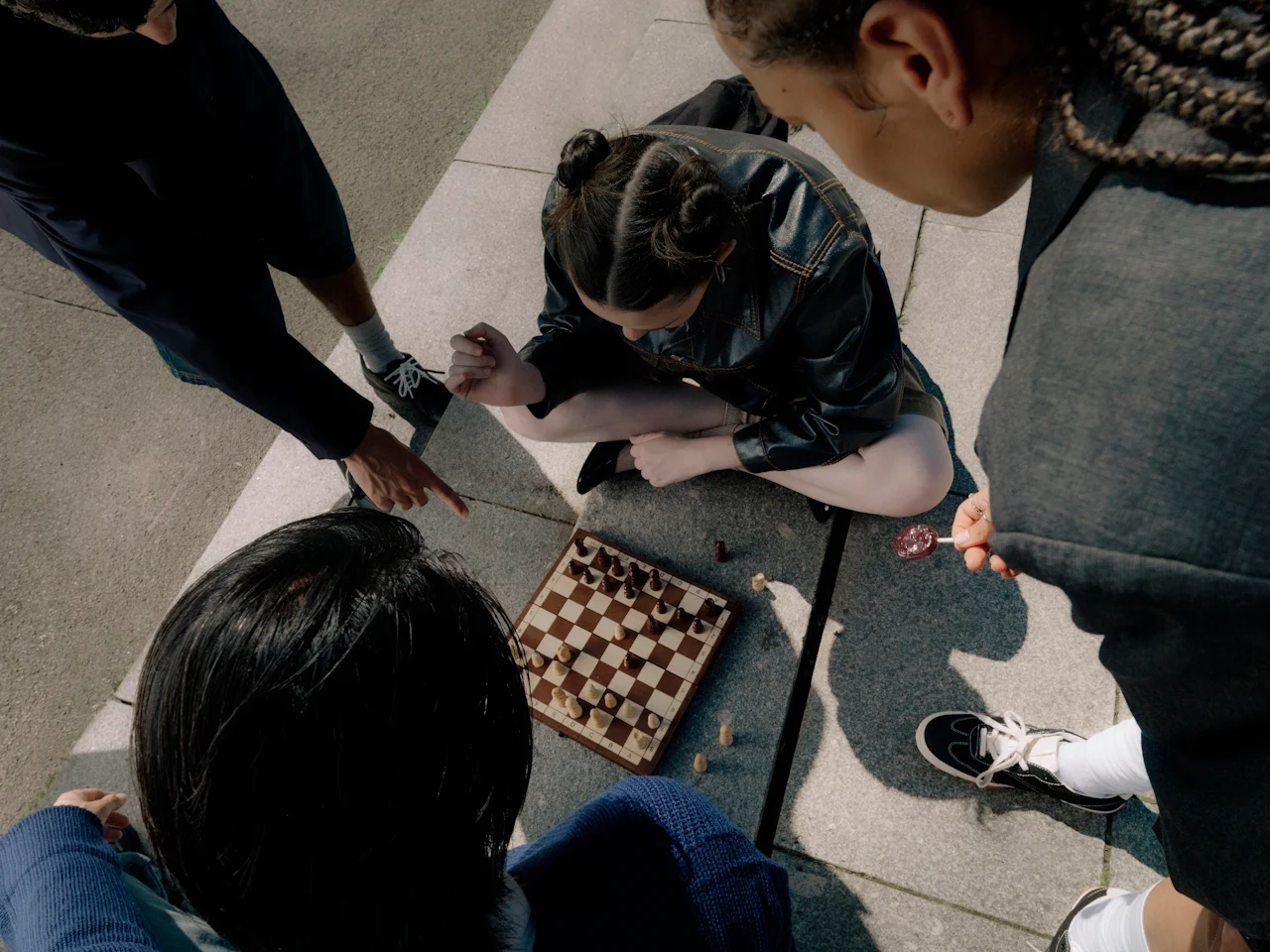 Four people are gathered outdoors, intently playing chess on a wooden board placed on the pavement. One person is sitting cross-legged, while others stand or crouch around the board, focused on the game.