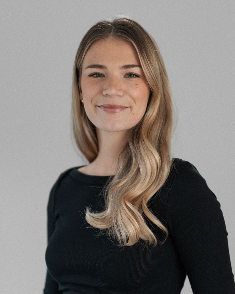 A young woman with long, wavy blonde hair and fair skin is wearing a black top and smiling gently at the camera against a plain light gray background, resembling Emily Dobson.