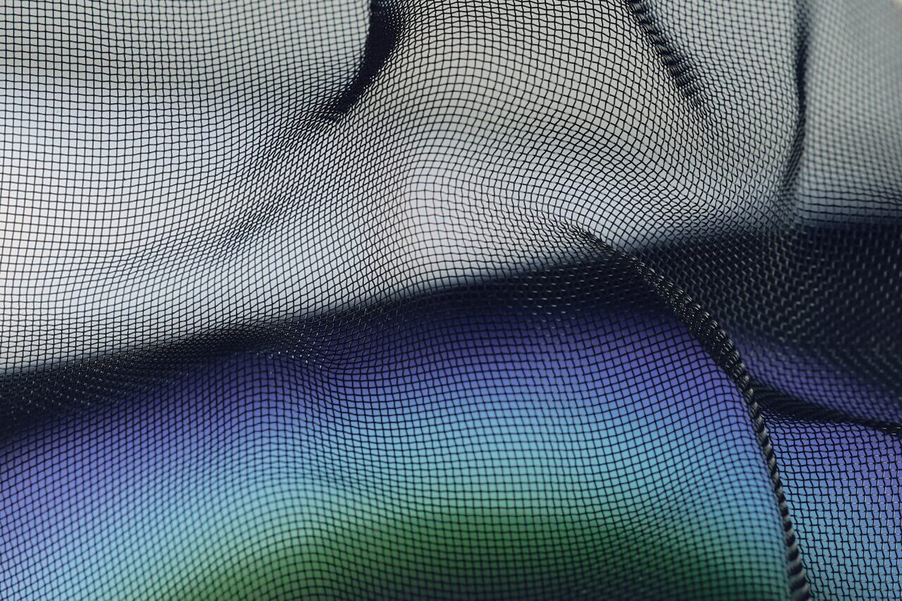 Close-up of a black mesh fabric with a grid pattern, draped in soft folds and shadows. Hints of blue and green blur beneath the mesh, hinting at a future-inspired, modern aesthetic.